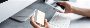 person sitting at a keyboard holding a bank card and a cell phone